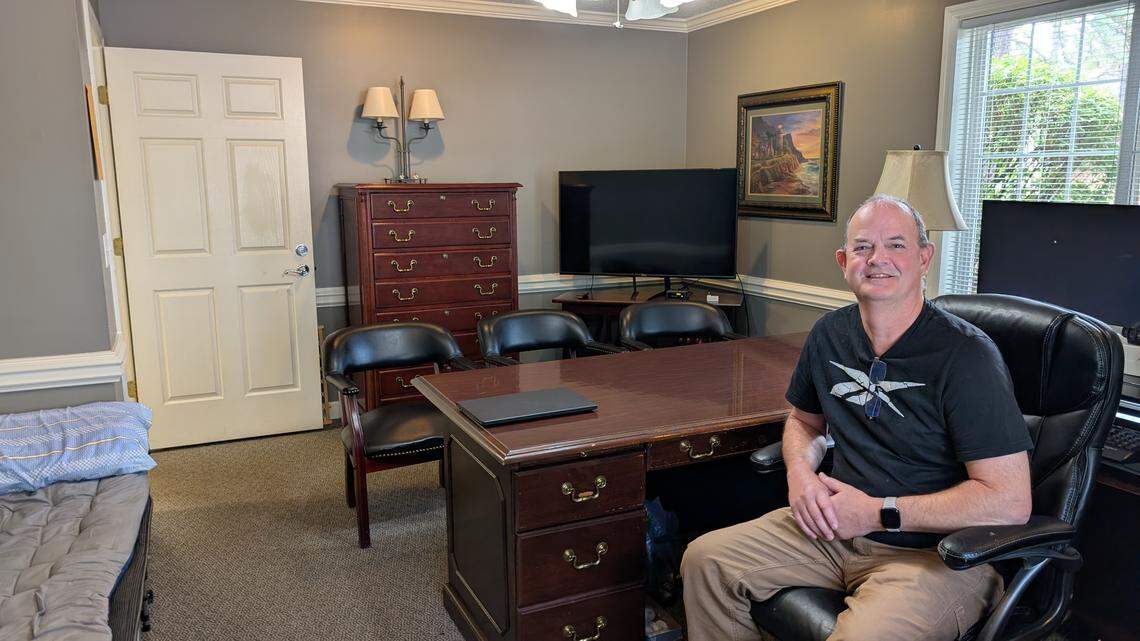 A full view of an office space. A person is sitting in a black leather chair behind a large wooden desk. The room has light gray walls, a closed white door on the left, a wooden dresser, a large television screen, and a window on the right. A cot with blue bedding is visible in the bottom left corner of the room.