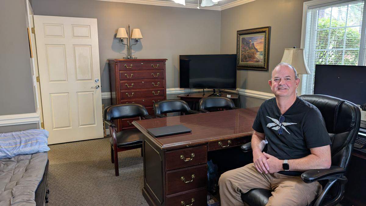 A full view of an office space. A person is sitting in a black leather chair behind a large wooden desk. The room has light gray walls, a closed white door on the left, a wooden dresser, a large television screen, and a window on the right. A cot with blue bedding is visible in the bottom left corner of the room.