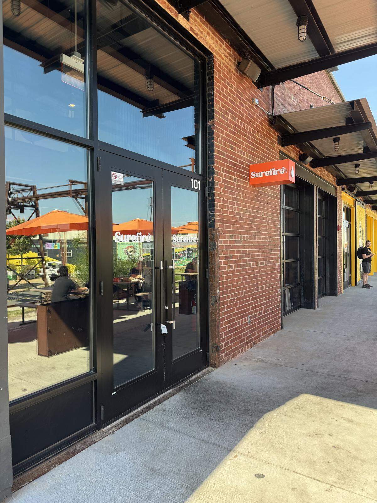 The brick exterior of a building with a large glass storefront and black doors with the Surefire Market logo visible on the glass and on a sign above the door. People can be seen sitting at tables with orange umbrellas outside, and another person stands near a yellow door in the distance.