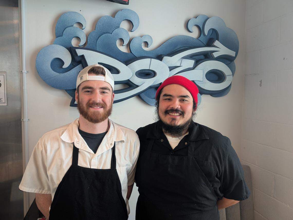 Two chefs smile while standing in front of a wall featuring a large, stylized logo with blue waves. The person on the left has a reddish beard and wears a white backward cap and black apron; the individual on the right has a dark beard and wears a red beanie and black apron.