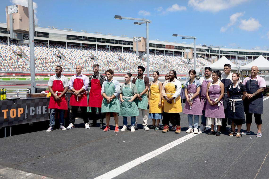 A group of contestants stands on the racetrack, some holding backpacks and bags, looking toward something off-camera. The massive grandstands of the Charlotte Motor Speedway tower behind them.