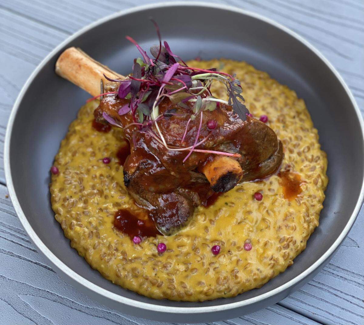 A braised lamb shank rests atop a bed of golden-yellow squash and farro risotto in a matte grey bowl. The lamb is coated in a rich, dark red reduction sauce and topped with a cluster of delicate purple microgreens. Small, bright pink peppercorns or garnish dots are scattered across the creamy surface of the grain. The dish is presented on a textured, grey wood-grain surface.
