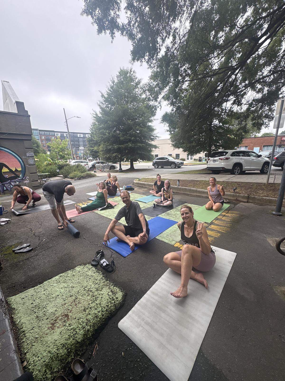 A slightly elevated outdoor shot captures a diverse group of about ten people engaged in a yoga class on a paved surface, likely a parking lot or street, under an overcast sky. Most individuals are seated on yoga mats in various poses. In the foreground, a woman in light purple shorts and a dark top is seated cross-legged on a light-colored mat, smiling and making a peace sign with her left hand. To her left, a man in a dark t-shirt and shorts sits on a blue mat, also smiling towards the camera. Further back, another man is bending over, seemingly rolling up a mat, while others are in seated or kneeling positions. Trees with green foliage line the background, and beyond them, cars are parked along a street. In the far background, urban buildings are visible, including one with a mural on its side to the far left. Green mossy patches are on the ground in the immediate foreground, and scattered shoes are near the mats.