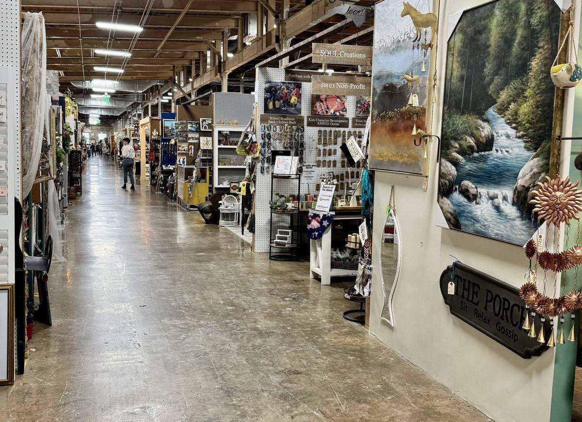 A perspective shot looking down a long, wide aisle in a large indoor artisans’ market. The industrial space is filled with individual booths on either side, displaying items for sale like paintings, signs, jewelry, and home decor. A few shoppers are visible in the distance.