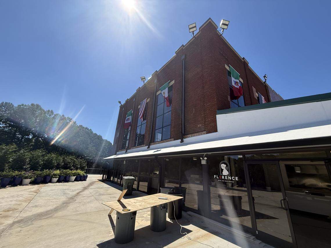 A wide-angle, sunlit view of a two-story brick building with large windows and an outdoor patio. Flags of Italy, Mexico, and other countries hang from the upper floor.