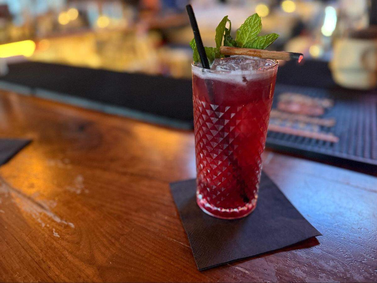 A close-up, eye-level shot of a deep-red cocktail in a tall, textured-glass, resting on a black napkin on a dark wooden bar top. The drink is garnished with a large sprig of fresh mint, a black straw, and a smoldering cinnamon stick. The background is a brightly lit but blurred bar.