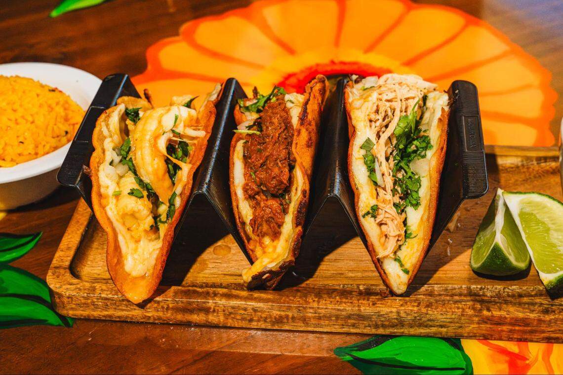 Three tacos filled with shredded meat and cilantro are presented in a black metal holder on a wooden platter. Fresh lime wedges and a side of yellow rice accompany the dish on a table painted with large orange and yellow flowers.