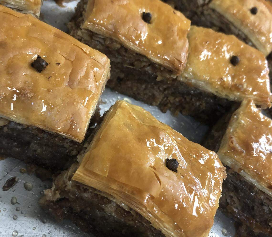 A detailed, close-up shot of homemade baklava. The diamond-shaped pieces feature a top layer of thin, golden phyllo dough and a thick middle layer of chopped nuts. The entire pastry is saturated with a glistening honey syrup, and each piece is garnished with a single clove.