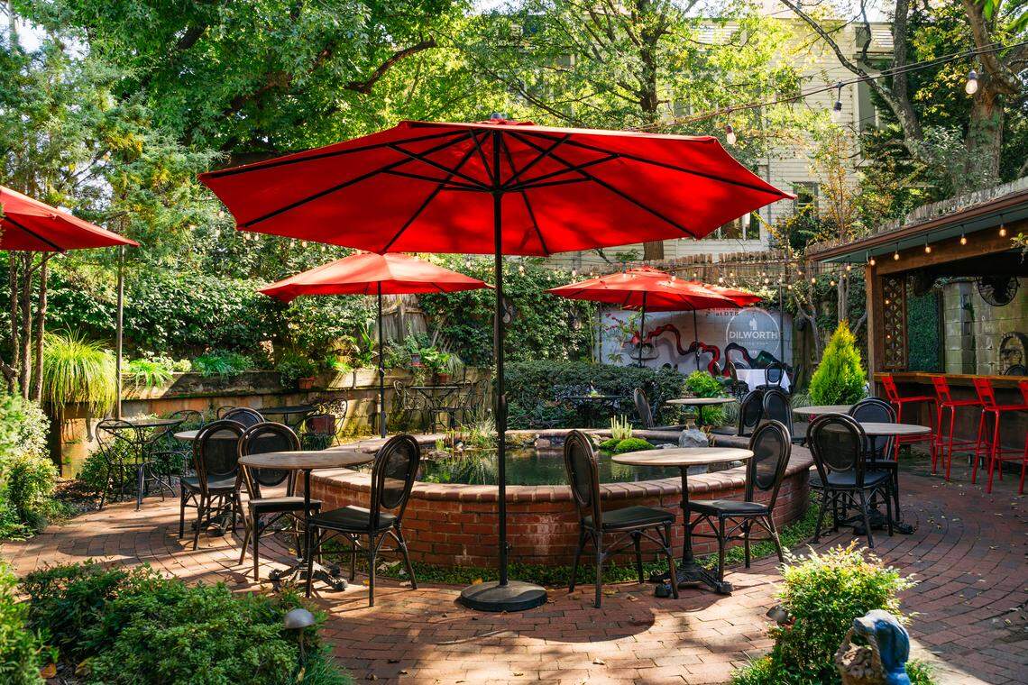 A picturesque, European-style garden patio is shown on a sunny day. The space, surrounded by dense trees and bushes, has a brick floor and a central brick-lined pond. Numerous tables are set for diners, with several large red umbrellas providing shade and a pop of color.