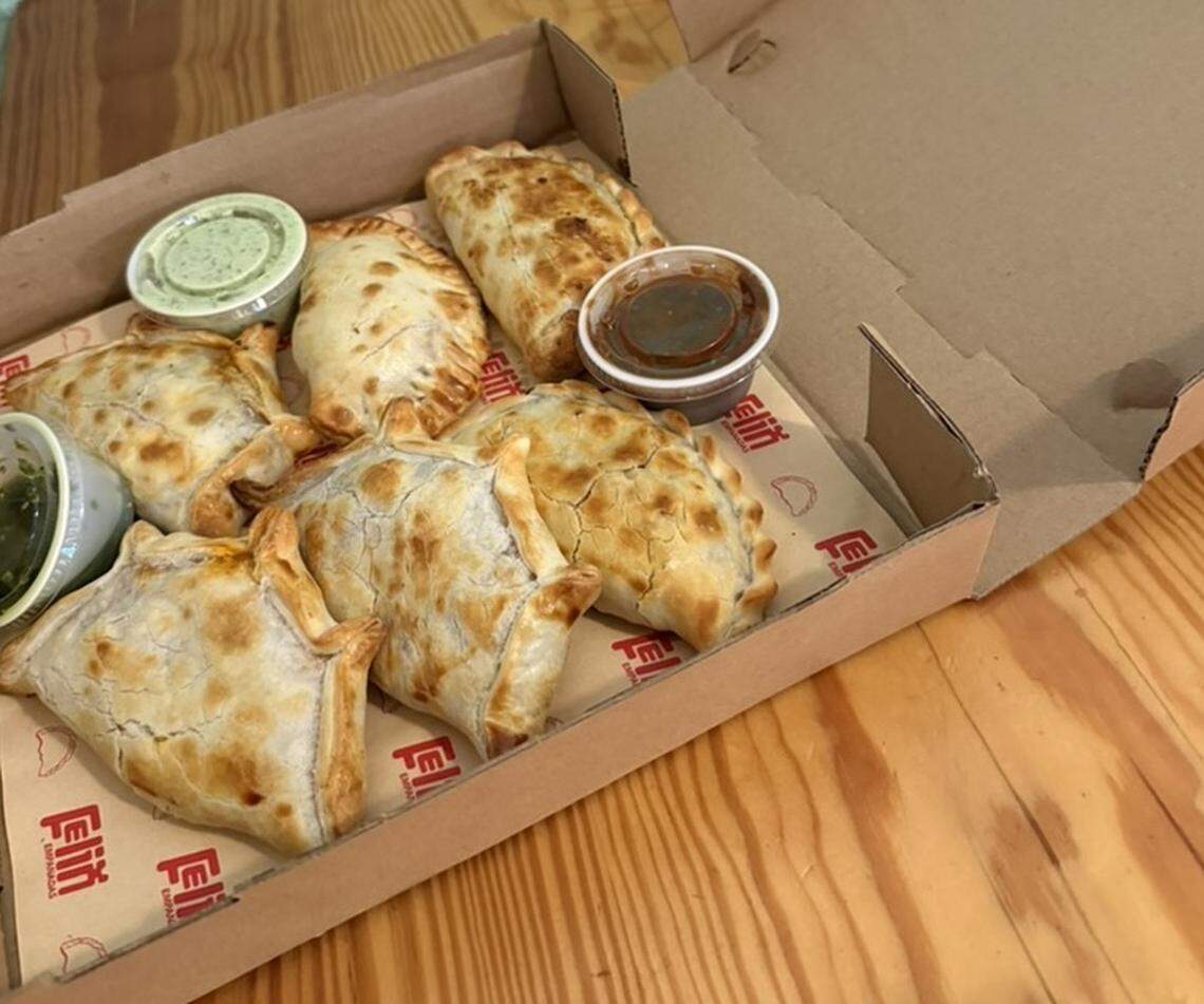 A cardboard takeout box sits open on a light wooden table, containing six golden-brown, baked empanadas. The empanadas have crimped edges and vary slightly in shape, resting on parchment paper branded with the red “Felix Empanadas” logo. Three small plastic containers of dipping sauces—two green (likely chimichurri or salsa verde) and one dark red—are tucked into the corners of the box.
