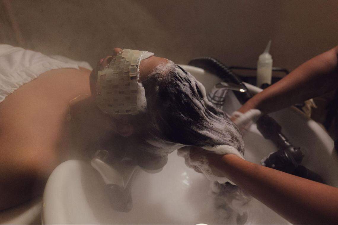 A close-up shot of a person receiving a luxurious hair wash. The client is wearing a cooling jade-style eye mask while a stylist’s hands massage shampoo into their hair. Steam fills the air, creating a relaxing atmosphere.