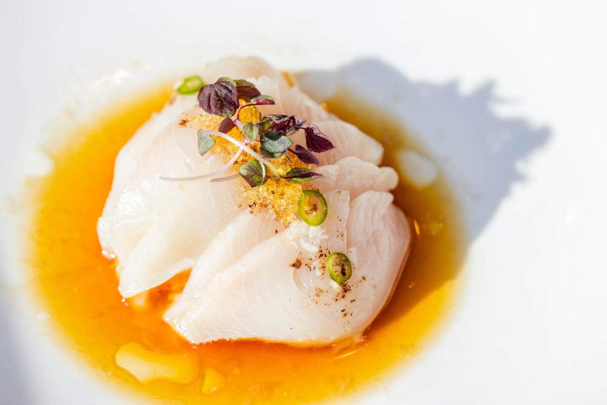 A high-angle, close-up shot of a delicate Japanese dish. Slices of pale, translucent hamachi (yellowtail) are neatly layered in a shallow white bowl containing a bright orange citrus-based sauce. The fish is garnished with golden pearls of fish roe, small purple microgreens, and thin slices of green serrano pepper.