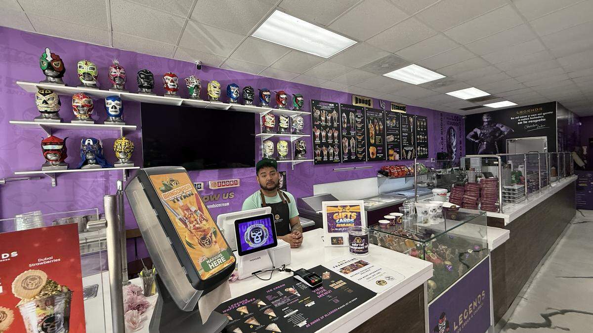 A high-angle, wide shot of the interior of a brightly lit shop with purple walls, taken from behind the service counter. An employee stands behind the counter, which holds a point-of-sale system. The back wall is decorated with multiple shelves displaying a large collection of colorful luchador masks, with a menu board and a large TV screen mounted below.