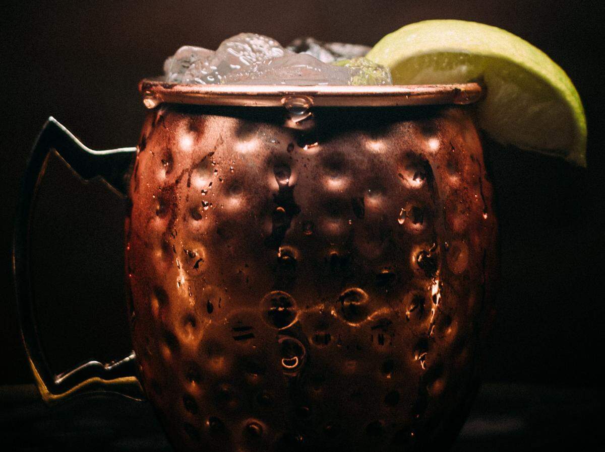A close-up shot of a cold Moscow Mule cocktail served in a classic, hammered copper mug. The mug is covered in condensation and has a bright lime wedge on the rim. The top of the drink is filled with crushed ice. The background is a dark, out-of-focus setting.