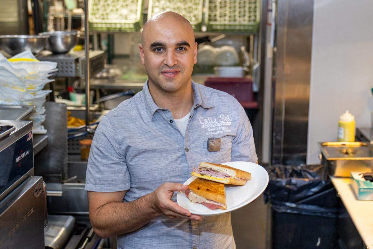 A medium shot of a bald chef, smiling and looking at the camera, wearing a gray “Calle Sol” executive chef shirt. The chef is standing in a commercial kitchen and holding up a white plate with a pressed Cuban sandwich, cut in half.