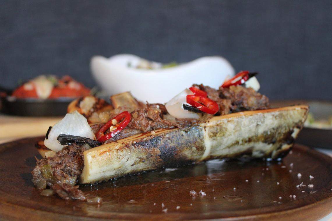 A close-up, side-angle shot of a large, halved bone filled with roasted marrow, topped with a generous portion of shredded meat. The dish is garnished with thin red chili rings, fresh white onion pieces, and is served on a dark wooden platter. A sprinkle of sea salt is visible on the platter. The background is blurred, showing other dishes on a table.