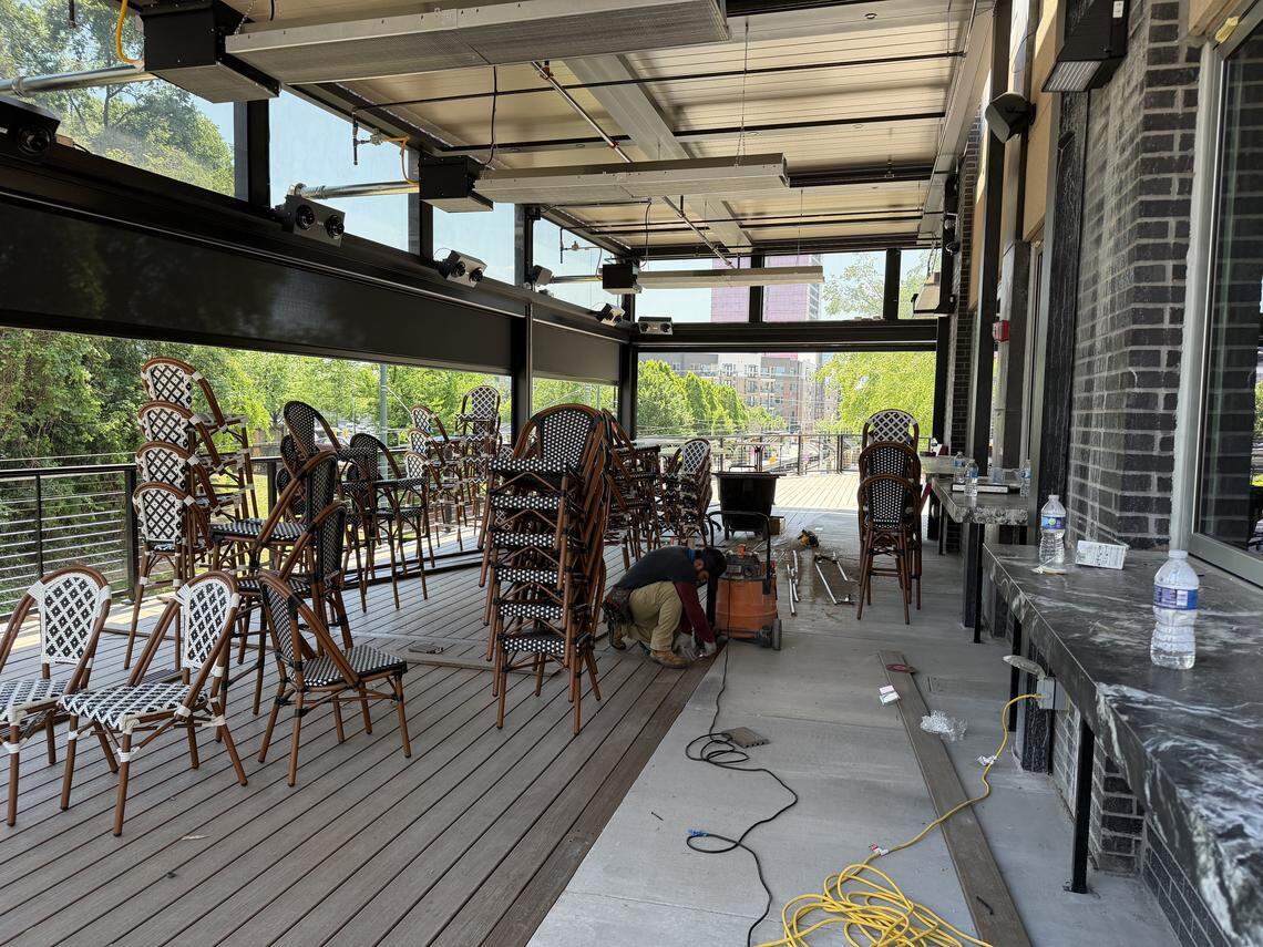 An covered outdoor patio under construction, featuring wooden decking and a long marble bar counter. A worker in a dark shirt and tan pants kneels on the floor near a shop vacuum and tools. Numerous bistro-style chairs with black-and-white woven patterns are stacked throughout the space, which overlooks green trees and distant city buildings.