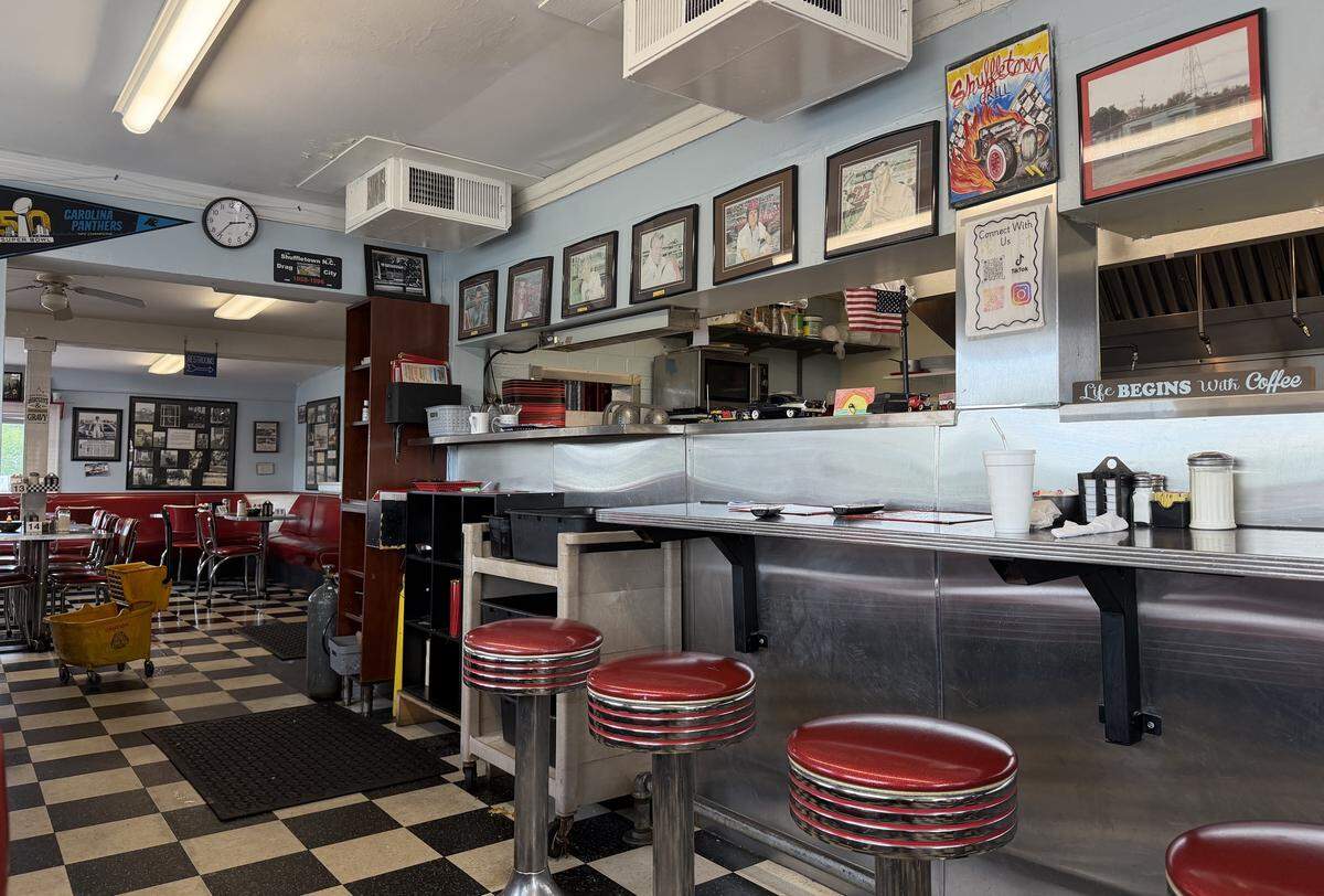 An interior view of a classic diner featuring a stainless steel counter with red vinyl swivel stools and a black-and-white checkered floor. The walls are decorated with framed sports photos, a Carolina Panthers pennant, and local memorabilia. In the background, a dining area with red booths and tables is visible. A sign above the counter reads, “Life Begins with Coffee.”