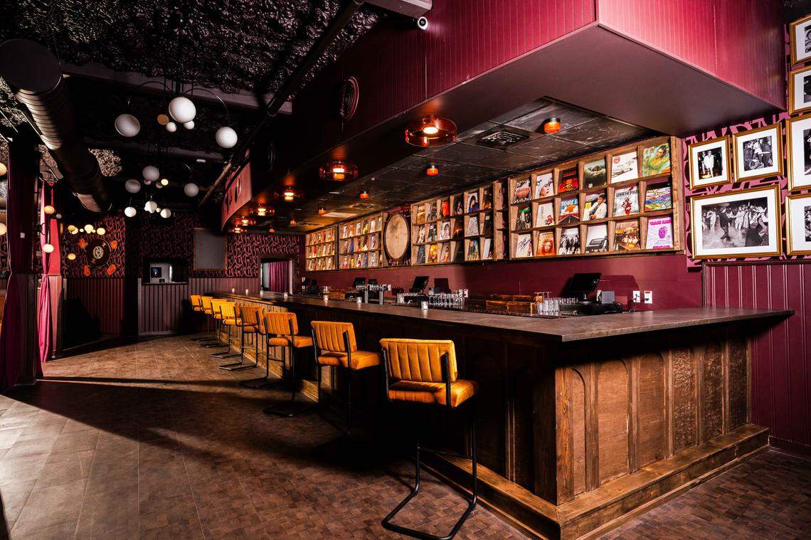 A wide shot of a sophisticated bar interior with a long, dark countertop and golden-yellow upholstered bar stools. The back bar is lined with multiple wooden shelves displaying a large collection of vinyl record covers. The room features deep red walls, a dark textured ceiling, and a mix of amber-tinted pendant lights and white globe lights, giving the space a moody, mid-century modern vibe.