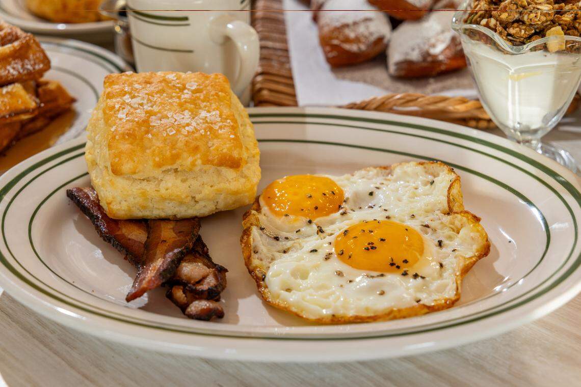 A breakfast plate featuring two sunny-side-up eggs seasoned with cracked black pepper, two slices of crispy bacon, and a large, golden-brown flaky biscuit topped with coarse sea salt. The food is served on a white diner plate with green rim stripes. In the blurred background, other breakfast items like beignets, waffles, and a yogurt parfait are visible.