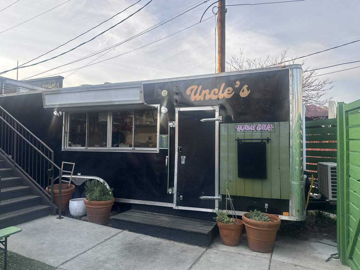 A wide shot of the Uncle’s food trailer, a sleek black mobile kitchen parked in an outdoor space. The trailer features a golden, cursive “Uncle’s” logo and a service window where the staff prepares orders. To the right, a green wooden partition displays a small sign that says “BURIAL GRUB.” Potted plants and herbs sit at the base of the trailer, and a set of metal stairs leads to an upper level in the background, creating a relaxed, urban patio atmosphere.