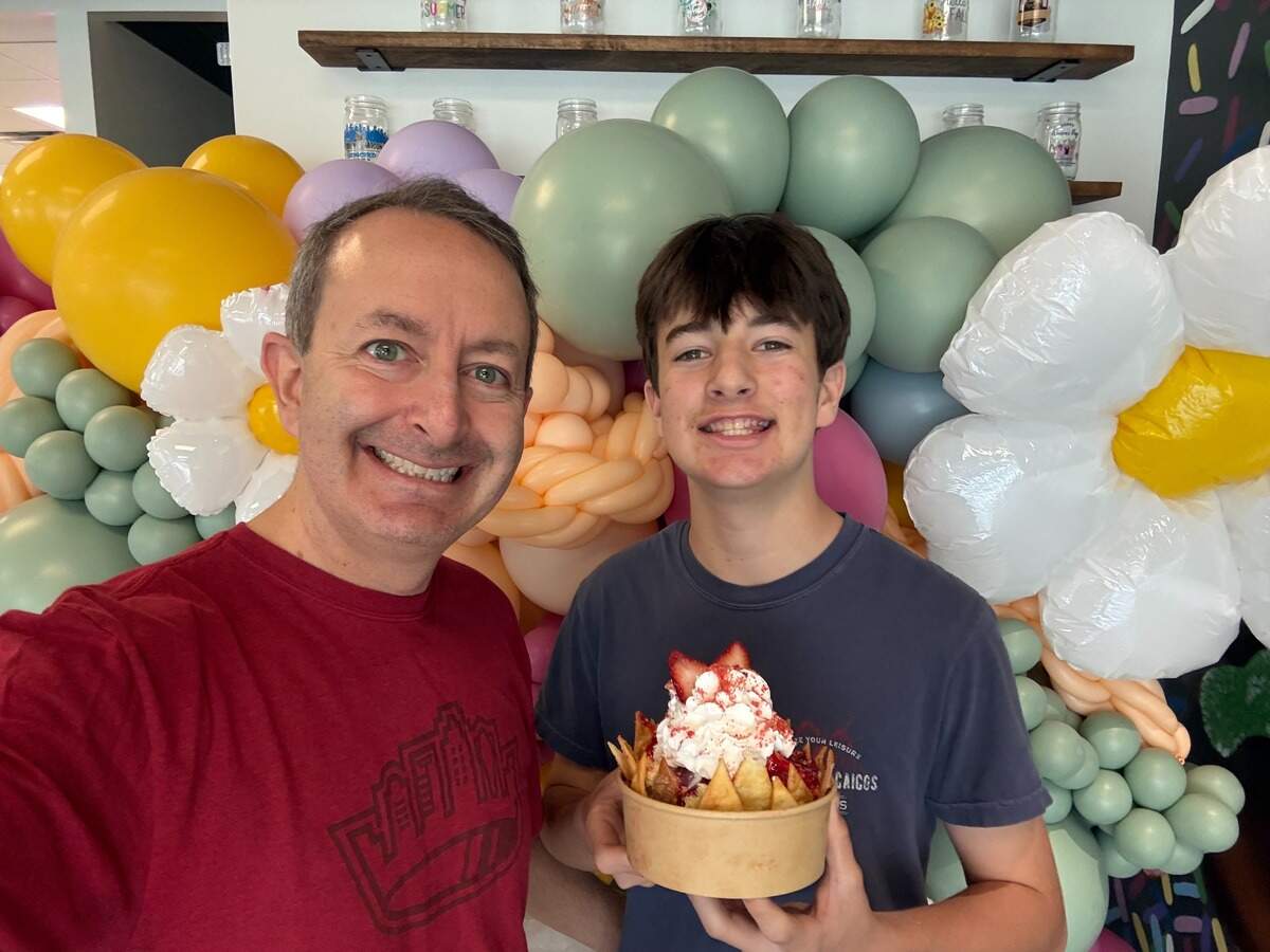 A father and child smile for a selfie while holding a strawberry dessert bowl in front of a colorful balloon arch.