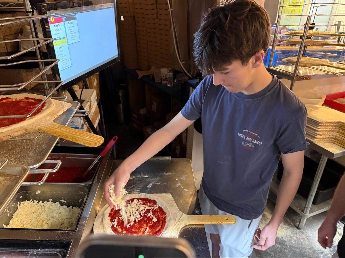 A high-angle shot of a young person at a prep station, carefully sprinkling shredded mozzarella cheese onto a raw pizza base spread with tomato sauce. To the left, a digital order screen displays incoming tickets.