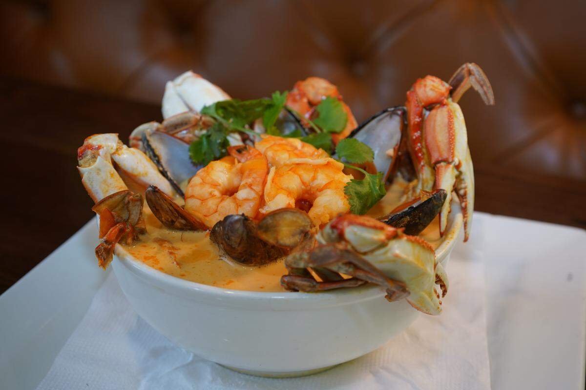 A luxurious and abundant bowl of seafood stew. A rich, creamy broth is packed with shellfish, including plump shrimp, dark-shelled mussels, and bright red crab claws and legs artfully arranged and spilling out of the bowl, all topped with a sprinkle of fresh cilantro.