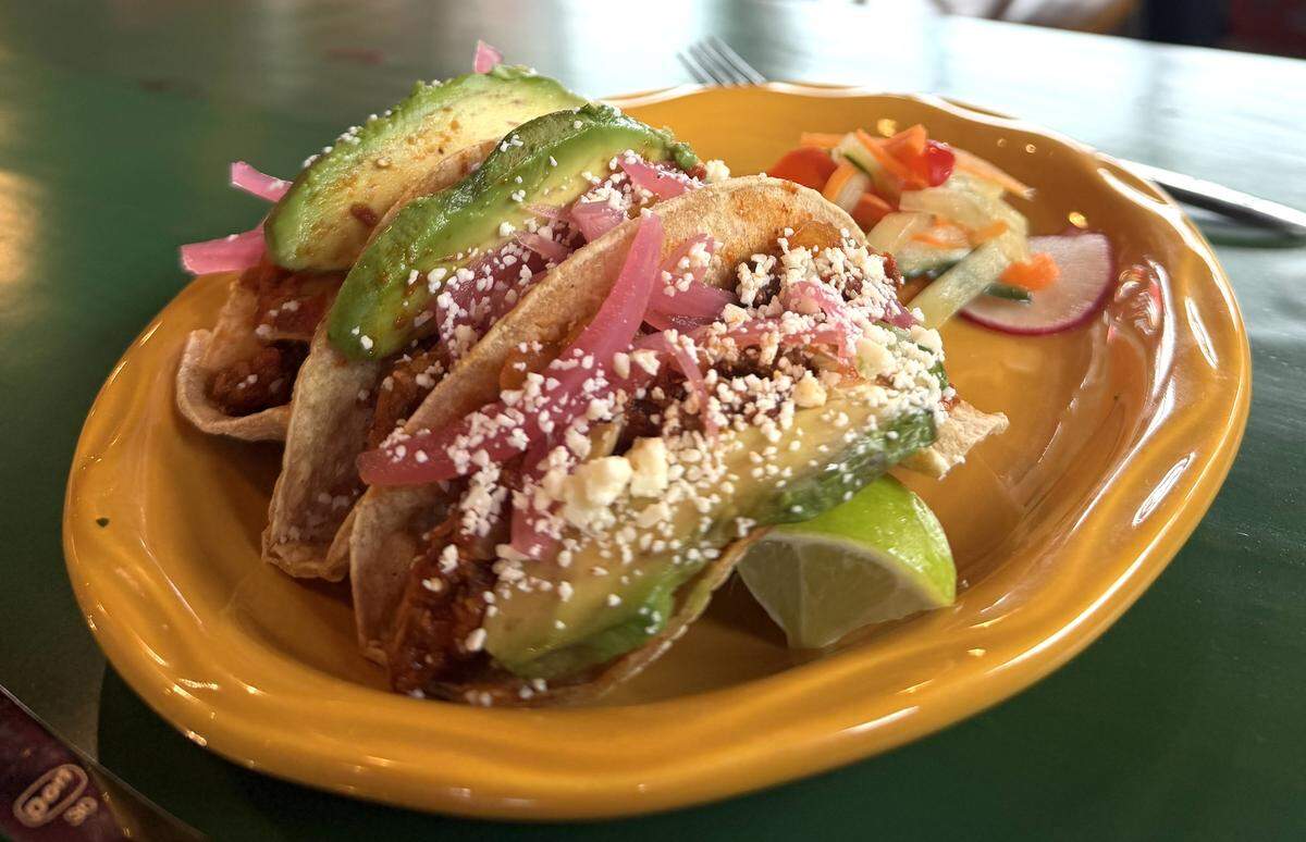 Three tacos in soft corn tortillas served on a yellow plate. The tacos are generously topped with thick slices of fresh avocado, pickled red onions, crumbled cotija cheese, and fresh cilantro. A small side of pickled vegetable slaw and a lime wedge are included.