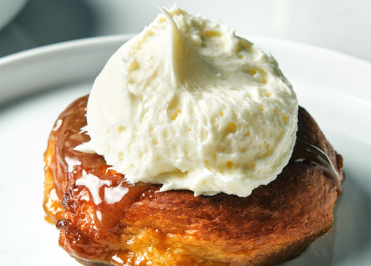 A close-up shot of a single, freshly baked sticky bun or pastry, topped with a generous swirl of light-colored frosting or whipped cream. The bun itself has a shiny, amber glaze, and its flaky texture is visible. It is served on a minimalist white plate, with the focus tight on the dessert.