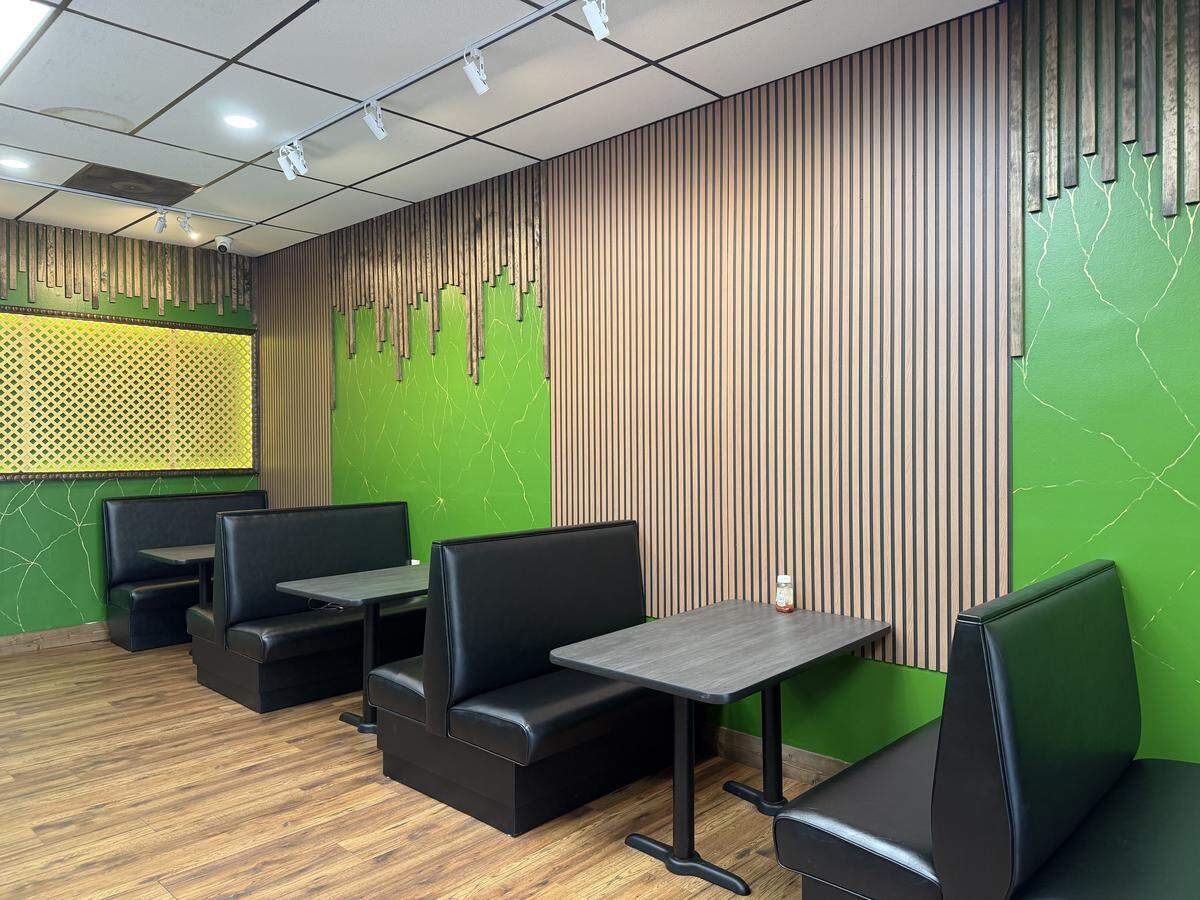 A wide, eye-level shot of a restaurant’s dining booth area featuring a row of four black vinyl booths paired with dark gray laminate tables. The distinctive accent wall behind the seating combines sections of vertical wood slats with vibrant green panels featuring subtle gold marble-like veining. In the background, a framed yellow lattice decorative piece is mounted on the wall. The space is lit by track and recessed ceiling lights, highlighting the warm wood-look flooring.
