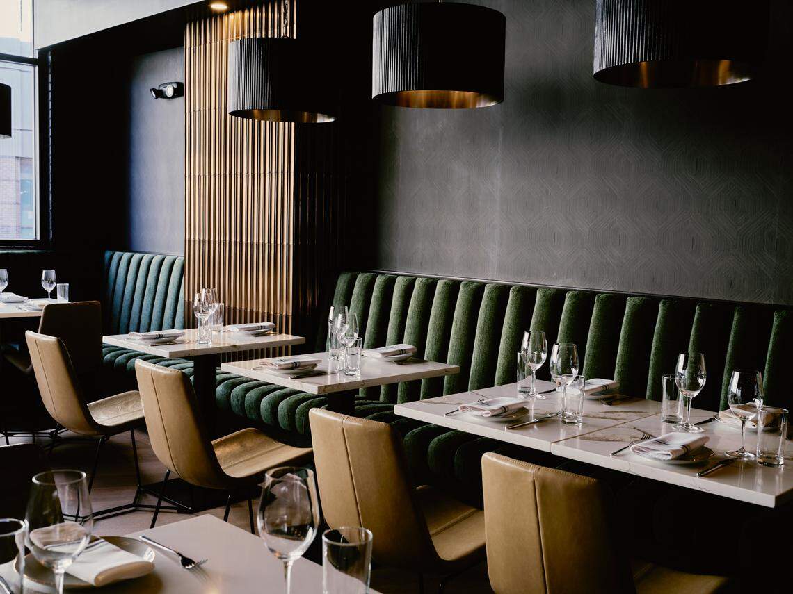 The interior of a dimly lit, modern restaurant. A long, dark green, channel-tufted banquette runs along a dark gray wall. In front of it are several tables with white marble tops, set with wine glasses. Tan leather chairs sit opposite the banquette. Large, black drum pendant lights with gold interiors hang from the ceiling, casting a warm glow.