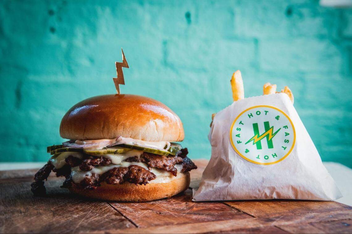A close-up, eye-level shot features a double cheeseburger and a bag of fries on a wooden cutting board against a textured turquoise wall. The burger consists of two beef patties, melted white cheese, pickles, onions, and sauce on a glossy brioche bun, topped with a gold lightning bolt-shaped pick. Next to it is a white paper bag of golden fries featuring a circular green and yellow logo that reads “HOT & FAST”.