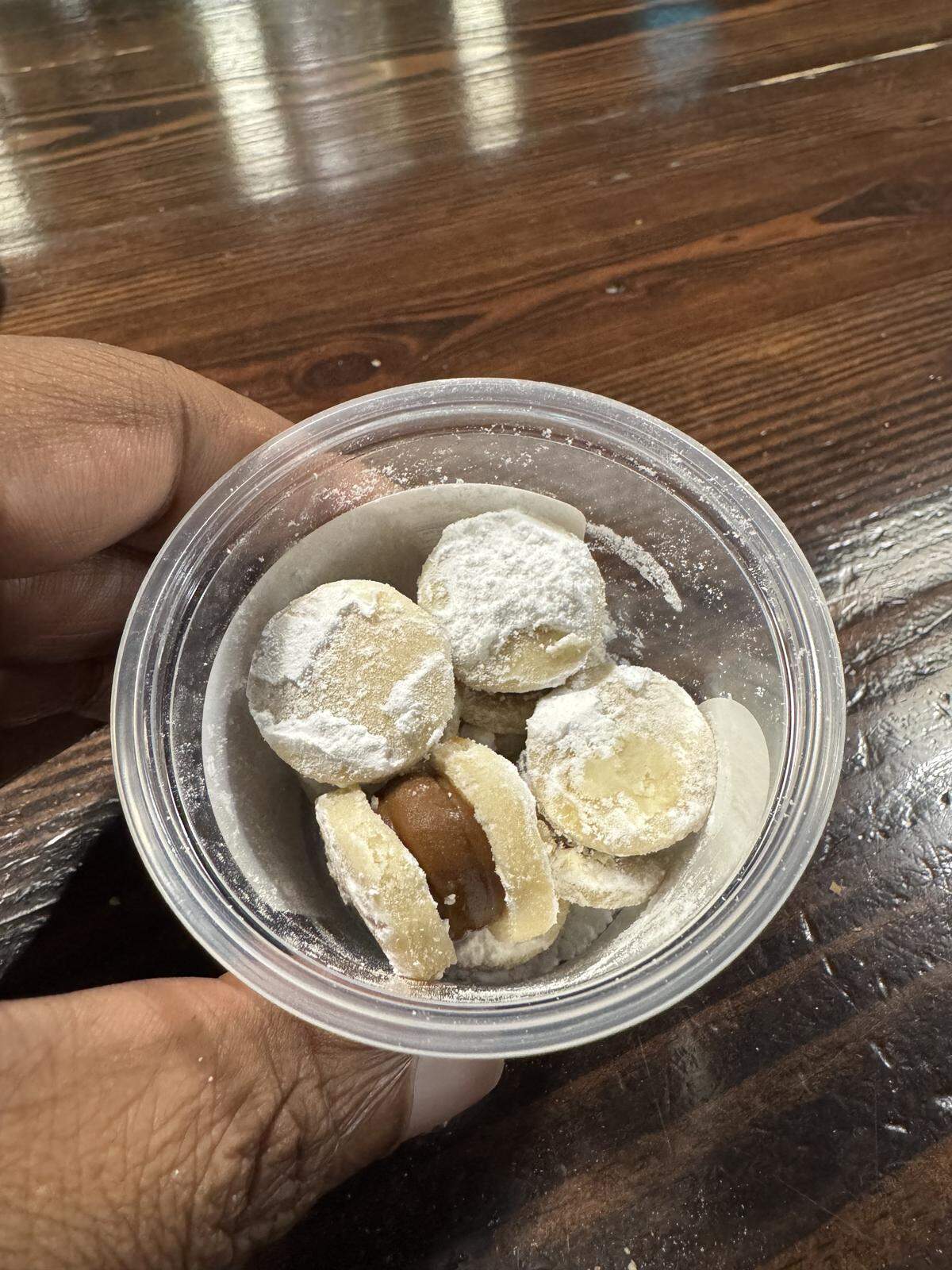 A close-up, top-down view of a small plastic container held by a hand, filled with several bite-sized shortbread cookies. The cookies are heavily dusted with powdered sugar, and one is shown as a sandwich cookie with a thick caramel or dulce de leche filling.