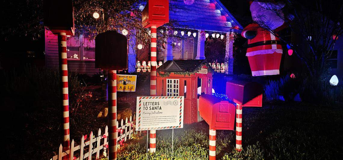 A front yard decorated at night with a whimsical Christmas display featuring multiple oversized red mailboxes on candy-cane striped posts, a small mailbox labeled ‘Letters to Santa,’ and a sign detailing instructions. The porch of the house behind the display is lit with blue and white lights.