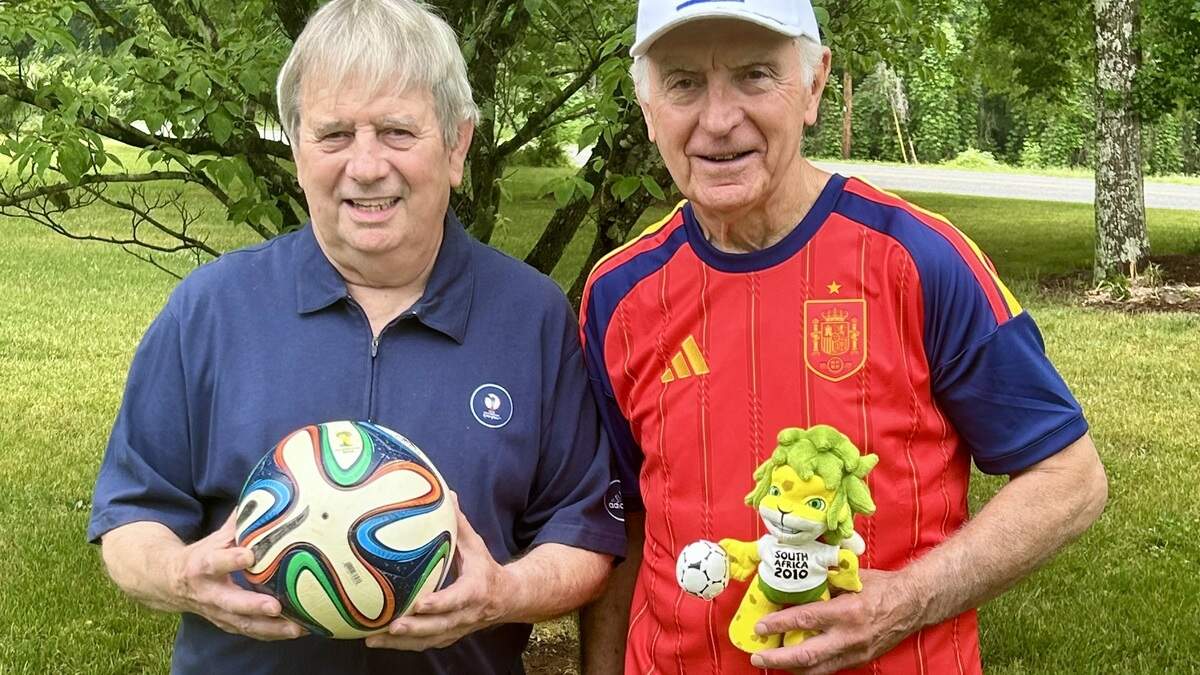 Terry Shinn (left) and Greg Crowley are longtime high school teachers who have made a hobby out of traveling to the World Cup every four years. Their first one was in 1994; the upcoming 2026 World Cup will be their ninth. They are holding souvenirs from previous World Cups: a soccer ball from the 2014 World Cup in Brazil and a stuffed toy from the 2010 World Cup in South Africa.