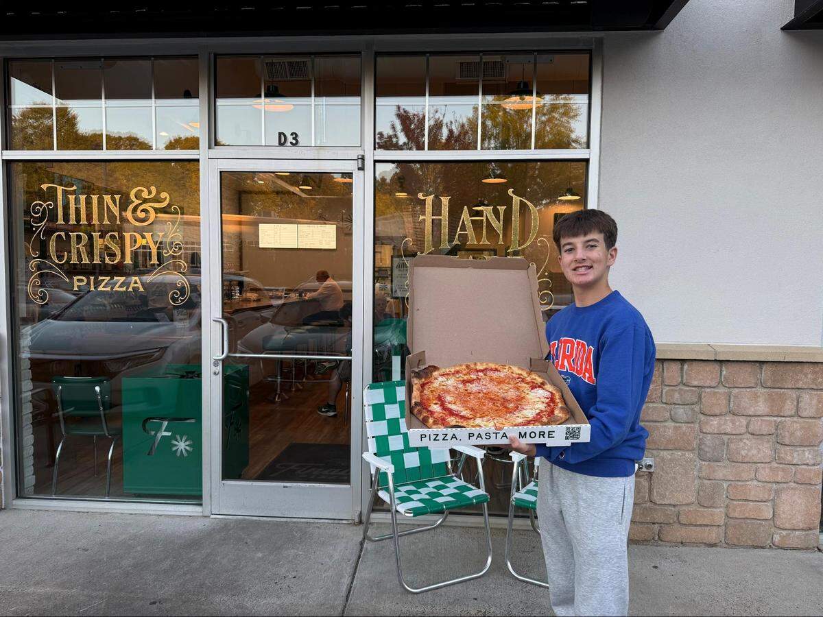 A medium, eye-level shot shows a smiling teenager standing outside a pizza restaurant, holding an open pizza box. The child, wearing a blue sweatshirt and grey sweatpants, holds up a “PIZZA, PASTA, MORE” box revealing a cheese pizza and standing on the sidewalk in front of the glass storefront, which has “THIN & CRISPY PIZZA” and “HAND” written on the windows.