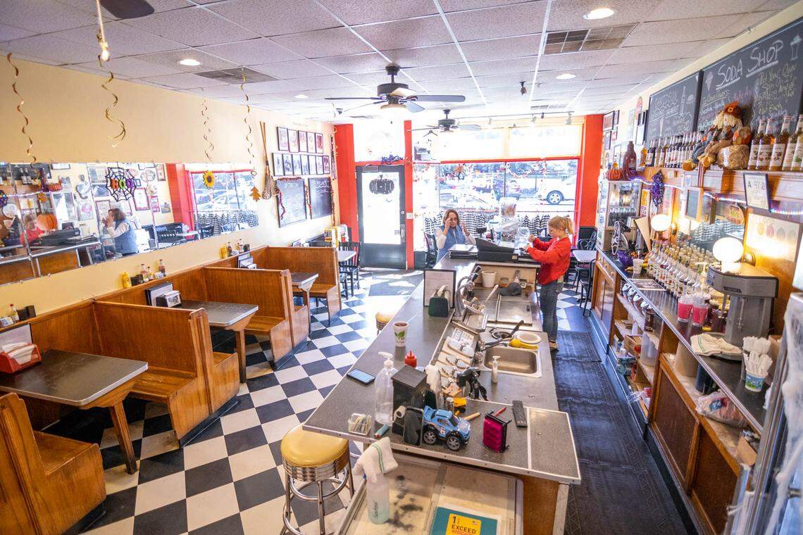 A high-angle, wide shot from the back of a retro-style diner called “The Soda Shop.” The restaurant features a black and white checkered floor, wooden booths along the left wall, and a long soda fountain counter in the center. A person in a red shirt, likely an employee, is working behind the counter, while a customer is seated at the counter near the front windows. The walls are decorated with framed photos and Halloween decorations, and a large chalkboard menu is visible on the back wall.