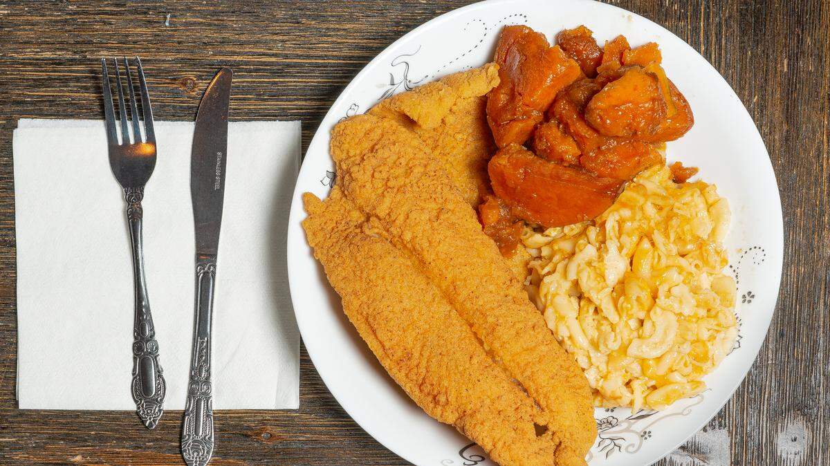 A top-down view of a white plate on a rustic wooden table. The plate contains two large, golden-brown cornmeal-crusted fried fish fillets, a serving of creamy baked macaroni and cheese, and a side of glazed candied yams. To the left, a silver fork and knife rest on a white paper napkin.