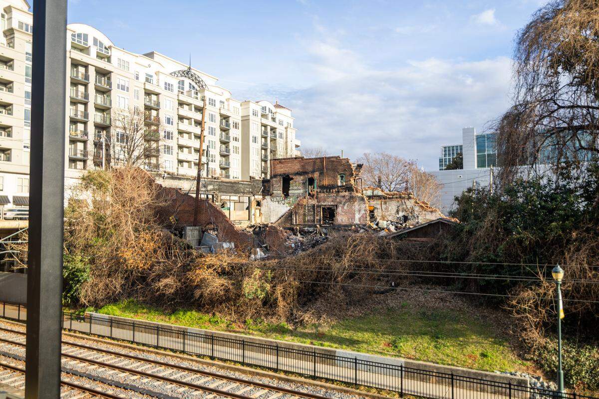 An outdoor view from an elevated position looking across light rail tracks toward the ruins of a historic brick building. In the background, a large, modern white apartment building stands in sharp contrast to the rubble and overgrown vines in the foreground.