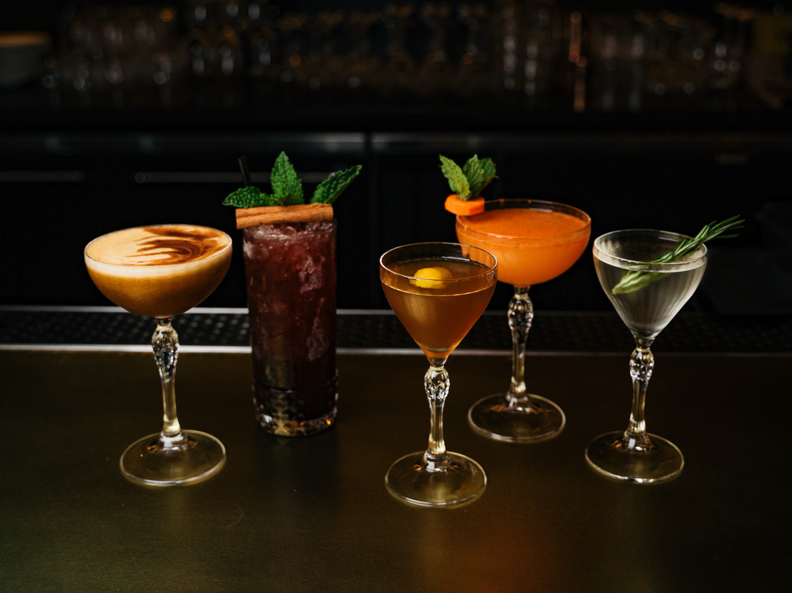 Five cocktail glasses are lined up along a dark wooden bar, each holding a different drink and garnish — cinnamon, mint, carrot and lemon.
