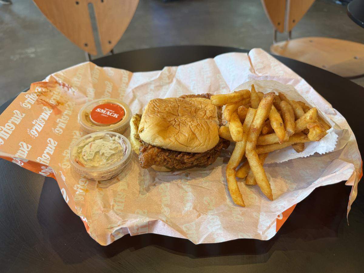 A fried chicken sandwich on a brioche bun with a side of French fries, coleslaw, and a small container of sauce sits on a piece of branded wax paper on a dark table.