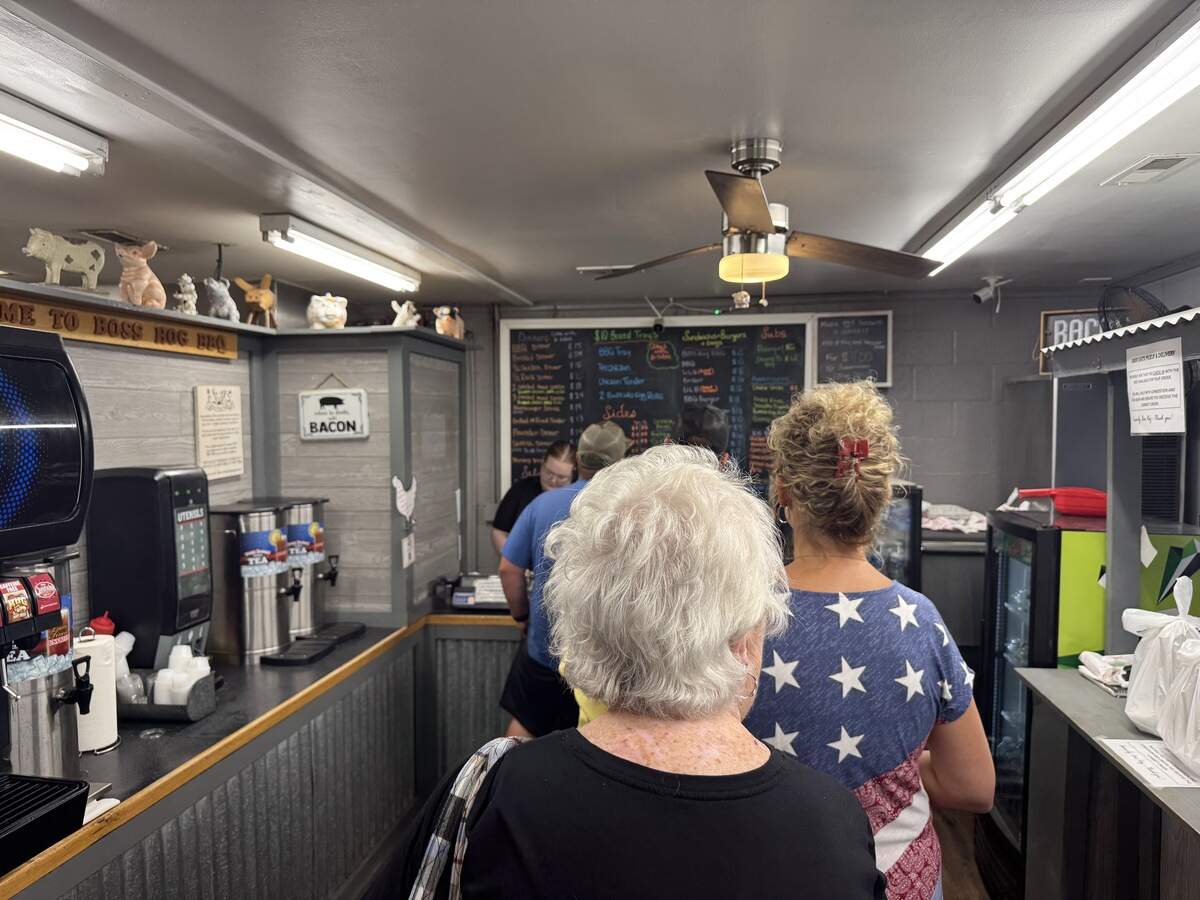 An indoor shot from the back of a line of customers waiting to order at the Boss Hog BBQ counter. A woman with short white hair stands in the foreground, followed by another woman wearing a patriotic blue shirt with white stars. In the background, a large black chalkboard menu hangs behind the counter where an employee is taking orders. To the left is a beverage station with sweet tea dispensers.
