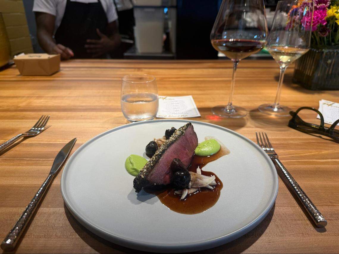 A wide shot of a chef’s counter featuring the seared protein dish from image 2. The wooden counter is also set with a glass of red wine, a small bowl of bread, a vase of yellow and purple flowers, and a pair of glasses.