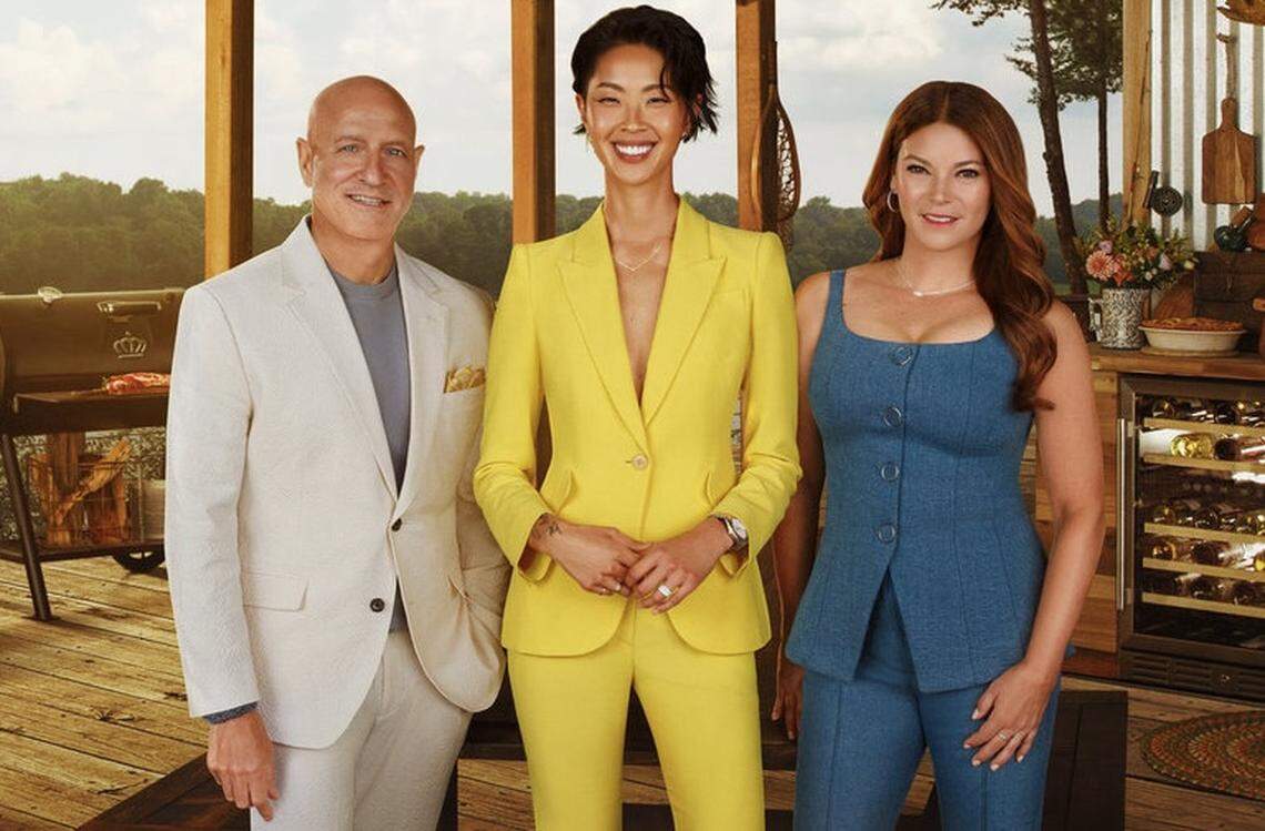 Three people standing together on an outdoor wooden deck with a scenic water and forest background. On the left, one in a cream-colored suit; in the center, one in a bright yellow tailored suit; and on the right, one in a blue denim-style top and matching pants. Behind them, an outdoor kitchen setup includes a grill, a wine refrigerator, and various kitchen tools.