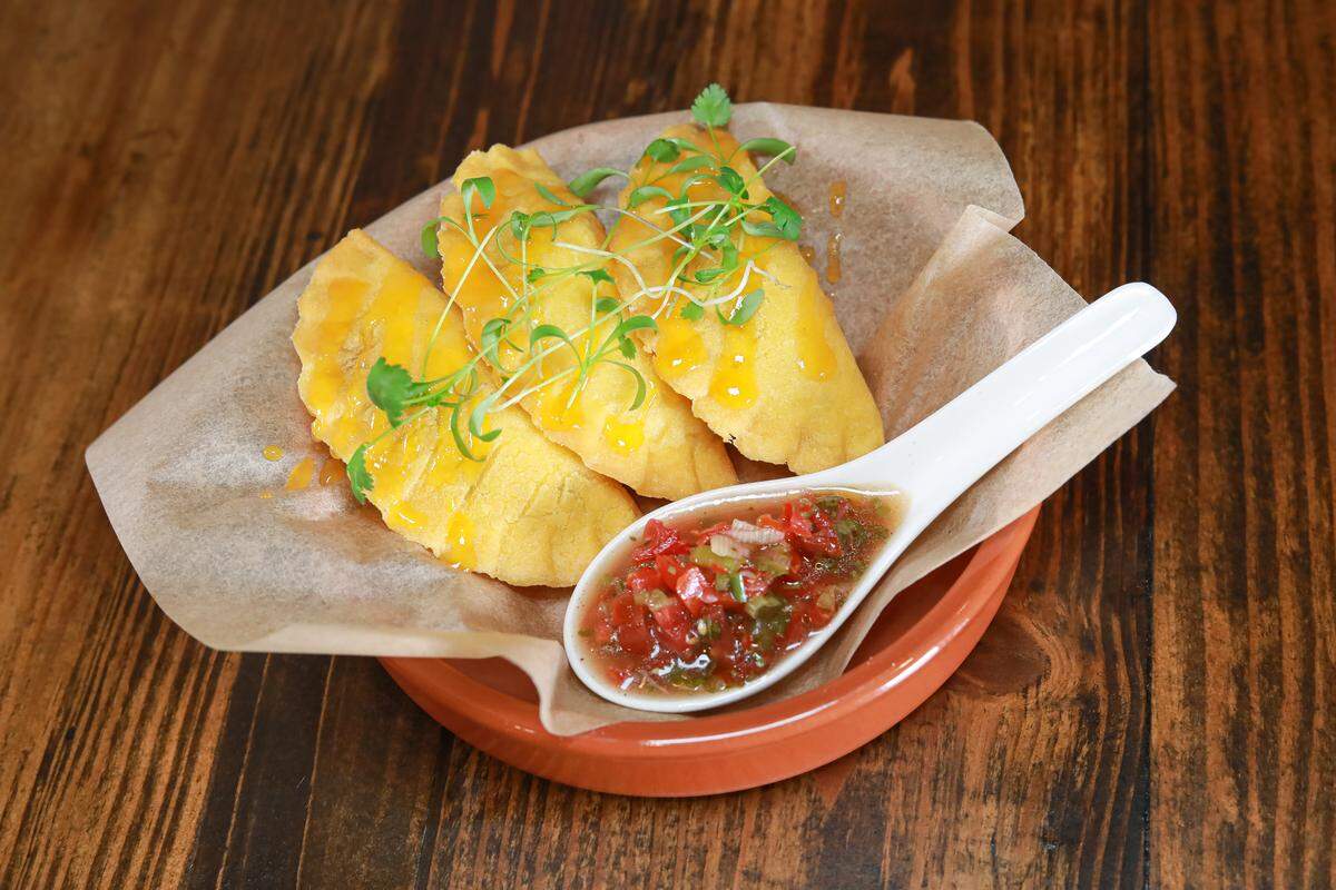 A high-angle, close-up shot of three golden-brown empanadas served in a terracotta dish lined with parchment paper. The empanadas are drizzled with a bright yellow sauce and topped with fresh microgreens. A white ceramic spoon filled with a chunky tomato and herb salsa sits in the foreground. The dish is placed on a dark, rustic wooden tabletop.