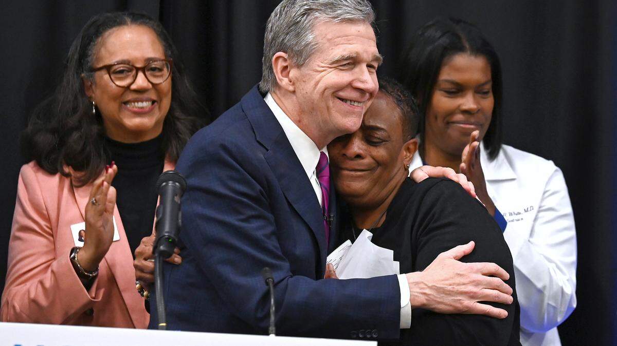 North Carolina Gov. Roy Cooper, left, hugs Penny Wingard, right, after presenting her with her new Medicaid card on Dec. 1, 2023. North Carolina became the 40th state to opt into the expanded Medicaid program. South Carolina is among the 10 remaining states that don’t participate.