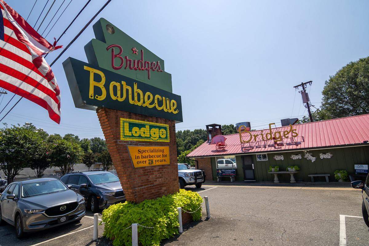 A low-angle, wide shot on a sunny day showing the exterior of Bridges Barbecue. In the foreground, a large sign with a brick base reads “Bridges Barbecue” in red and yellow text, with a smaller yellow “Lodge” sign below it. The restaurant, a single-story green building with a red roof, sits in the background. Cars are parked in the asphalt lot, and an American flag hangs from a pole on the left.