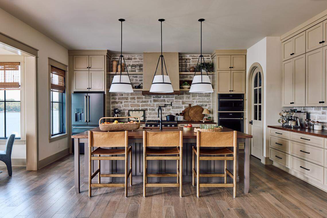 A modern farmhouse-style kitchen featuring light beige upper and lower cabinets and medium-toned wood flooring. The large central island has a dark wood top, a black sink faucet, and is flanked by three leather-seated, backless wooden bar stools. Above the island, three pendant lights with large white cone shades hang from the ceiling. The main wall features a backsplash of light, distressed brick behind the stovetop, which is covered by a tan-colored hood vent. Appliances include a dark, stainless steel refrigerator on the left and a double wall oven unit on the right. A tall, arched doorway is visible on the right wall.