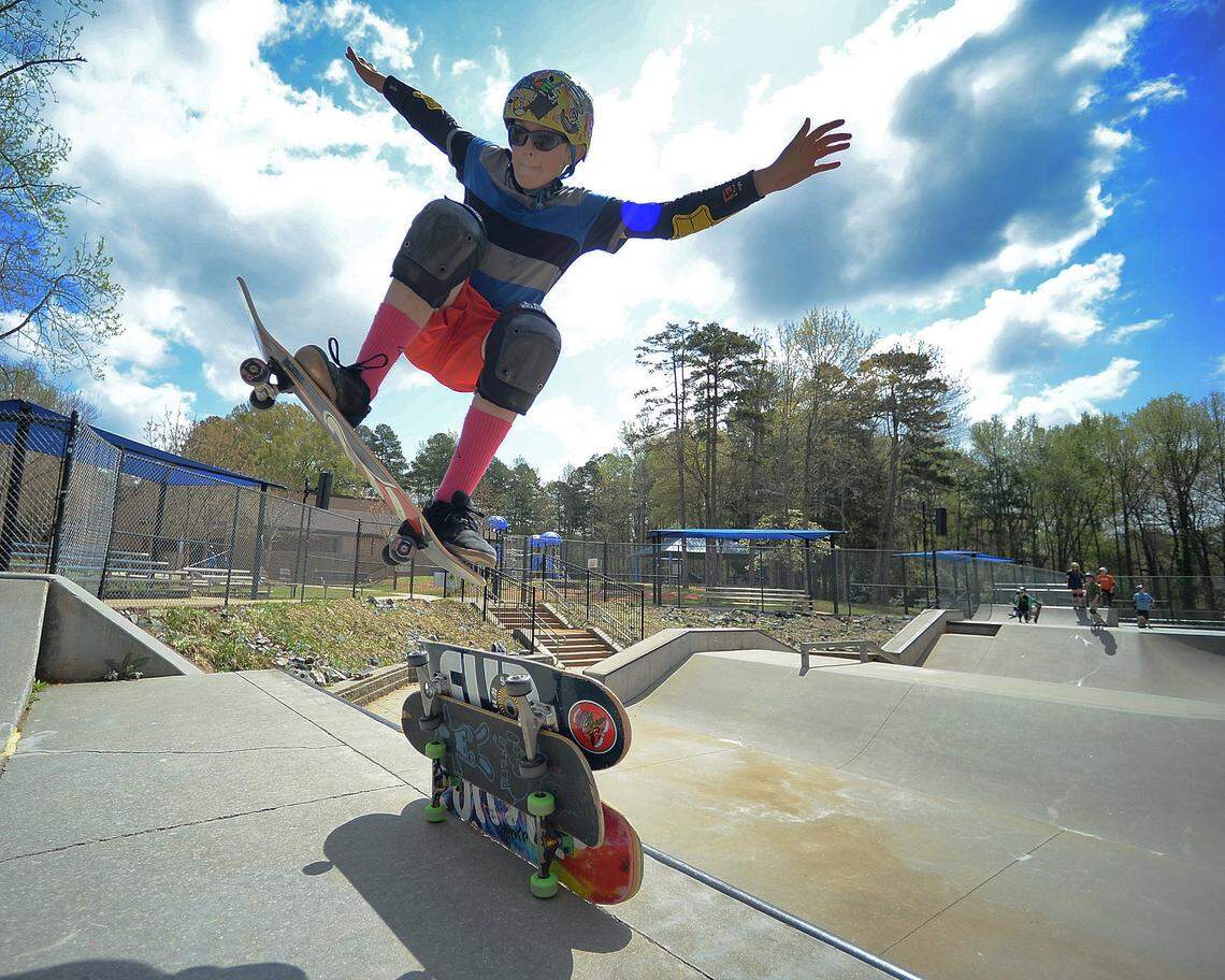 A dynamic, low-angle shot of a young person in mid-air on a skateboard at a skate park. The skater, wearing a helmet, sunglasses, and full protective pads, has their arms outstretched for balance against a bright blue sky with dramatic clouds. Two skateboards rest in the foreground, and the concrete ramps and bowls of the park, along with other skaters, are visible in the background.